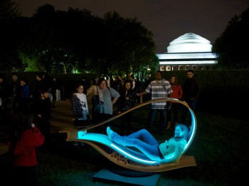 A person sits in a blue teardrop-shaped illuminated seat outside MIT's great dome while others look on.