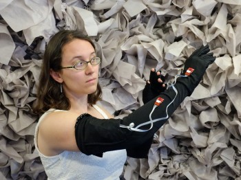 A woman wears long black gloves with wires on them in front of a wall of crumpled paper.