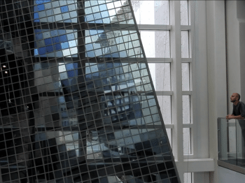 A person on a balcony looks at a large reflective sculpture made of many squares of glass.