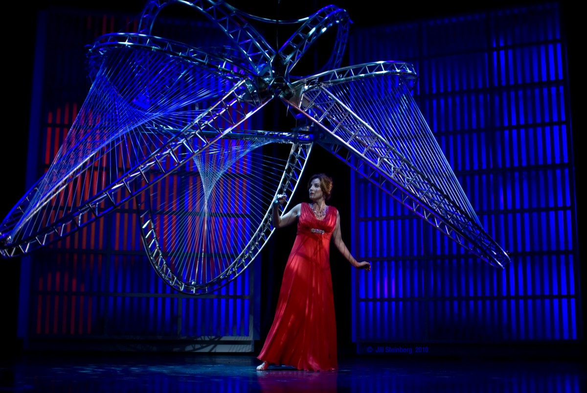 A woman in a red dress performs under a complex suspended metal structure.
