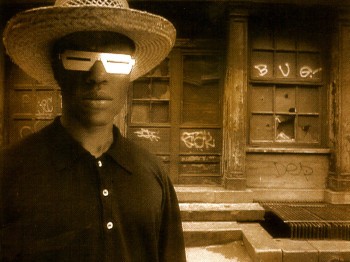 A man with futuristic eyewear stands in front of a dilapidated building.