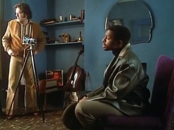 Video still of two men in a living room. One man has a camera on a tripod.