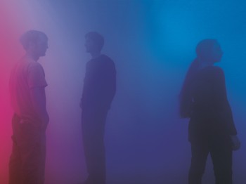 Three people stand in a dark space illuminated by low blue and purple light
