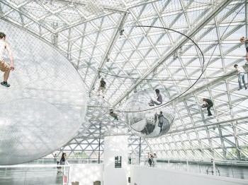People walk and sit on large nets suspended under a glass ceiling in a large atrium.