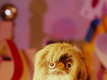 Video still of a wood puppet with lots of shaggy blonde hair.