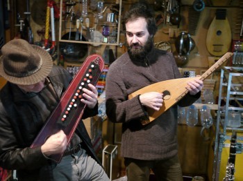 Two men play small stringed instruments in an instrument workshop.