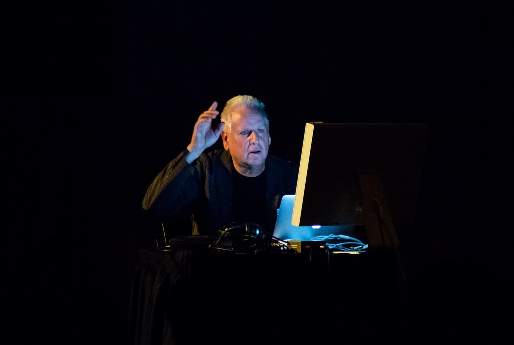 A man sits at a table with a laptop and a monitor in a dark room.