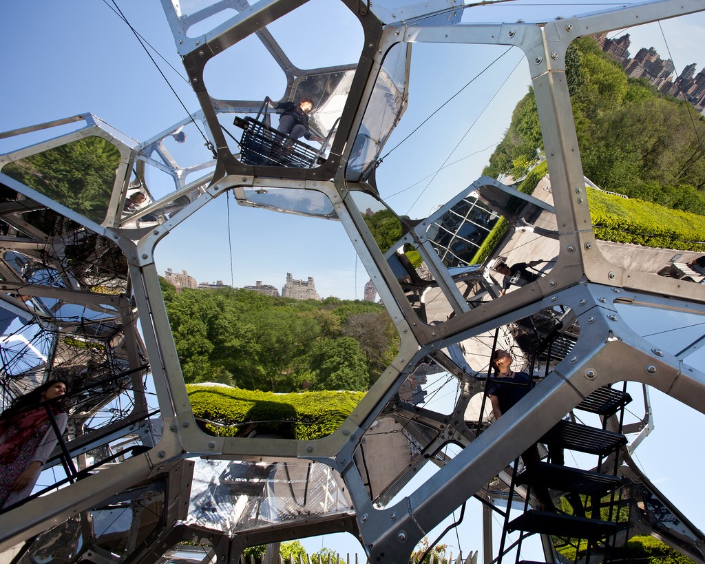 A structure made of interlocking geometric mirrors and panes of glass, reflecting buildings and trees.