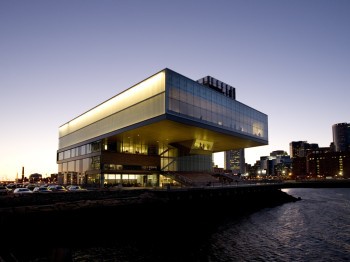 A large cube-shaped building with a top floor that projects out. The building sits on a waterfront.
