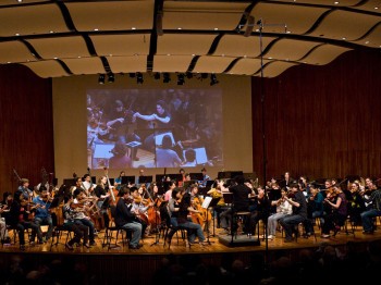Musicians perform on a stage with a live projection of the conductor behind them