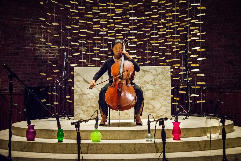 A woman performs cello surrounded by glass vases and microphones.