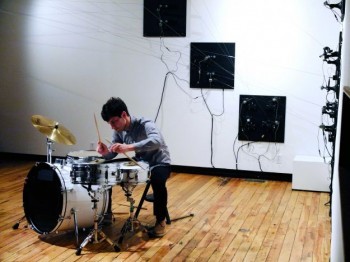 A man plays a drum kit in a room crisscrossed with many wires connected to panels on the walls.
