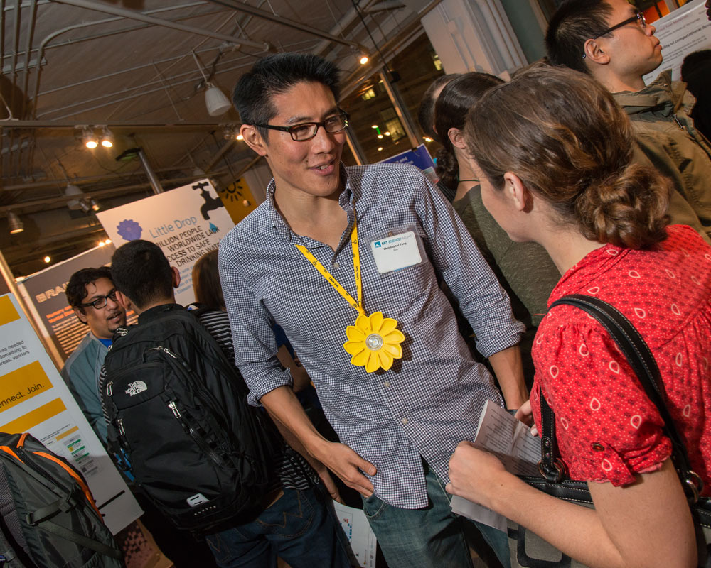 A man wearing a sun-shaped light on a lanyard speaks to a person at a busy event.
