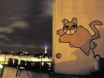 Graffiti of a smiling cat on a cement wall. The Eiffel Tower is illuminated in the background.