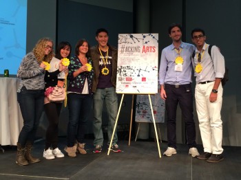 Four people wearing sun-shaped lights on lanyards pose next to a sign reading "Hacking Arts"