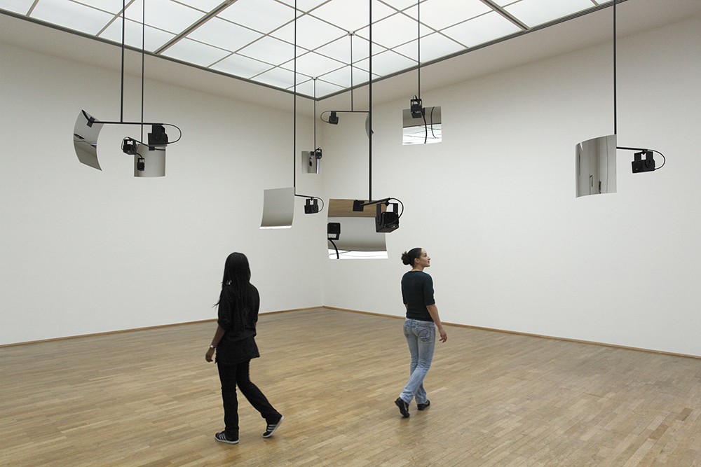 Two people in a gallery with installations of curved mirrors and electronic devices suspended from the ceiling