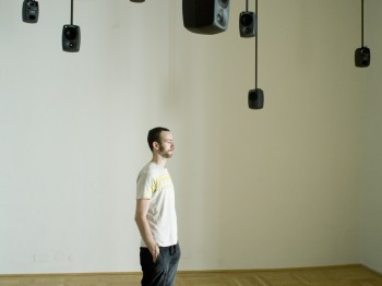 A man stands among speakers suspended from the ceiling.