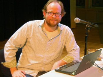 A man poses next to a microphone and a laptop.
