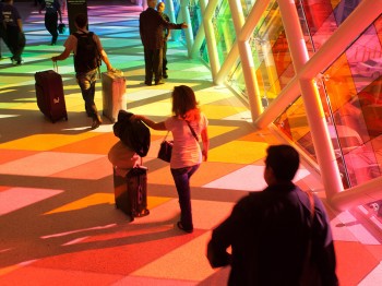 People walk along a hallway illuminated by sunlight coming through multi-colored panes of glass.