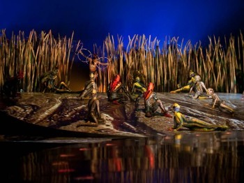 Performers in lizard-like costumes on a stage set depicting a river's edge