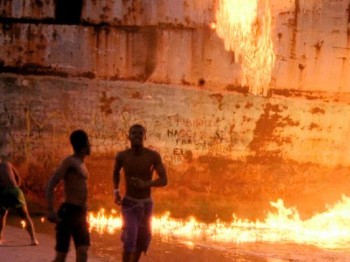 Three people in front of a large rusted ship and flames in the water next to it.
