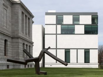 A geometric metal sculpture on a lawn outside a grey building and a white building.