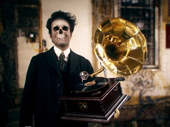 A man whose face is a skull holds an old phonograph in a dilapidated room