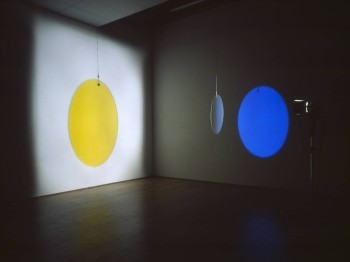 Art gallery showing a hanging disc, which projects a yellow image on one wall and a blue image on another wall when illuminated by a spotlight.