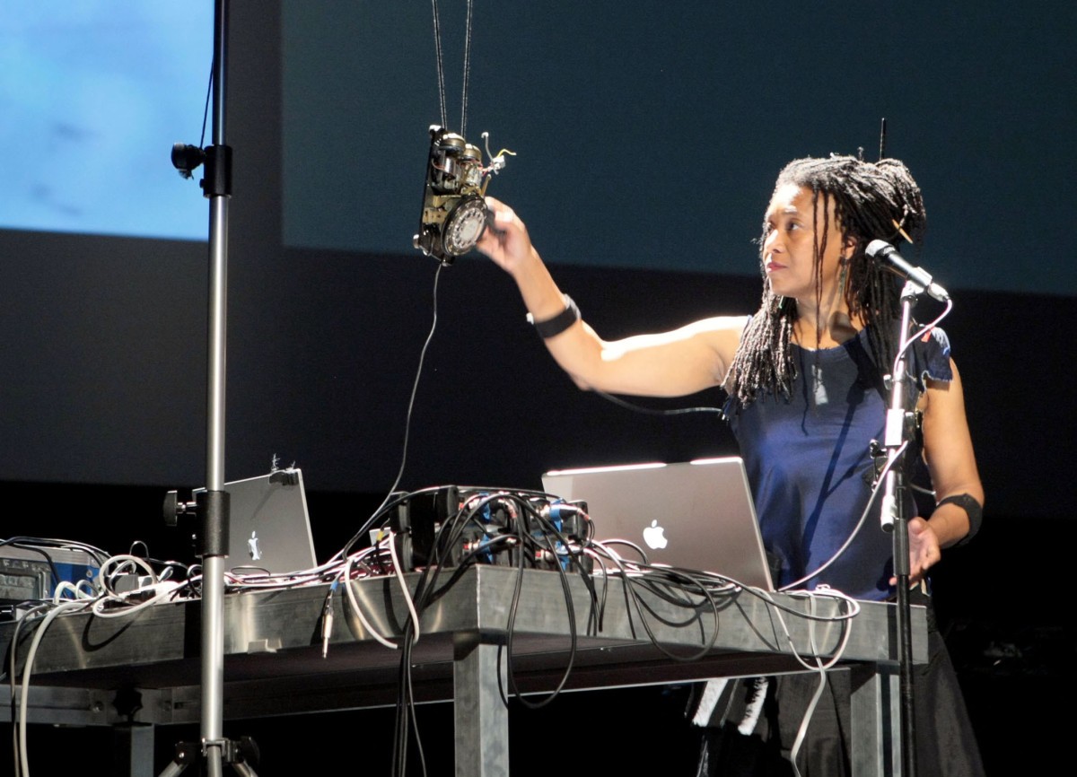 A woman on a stage stands next to a microphone and works with laptops and electronic equipment.