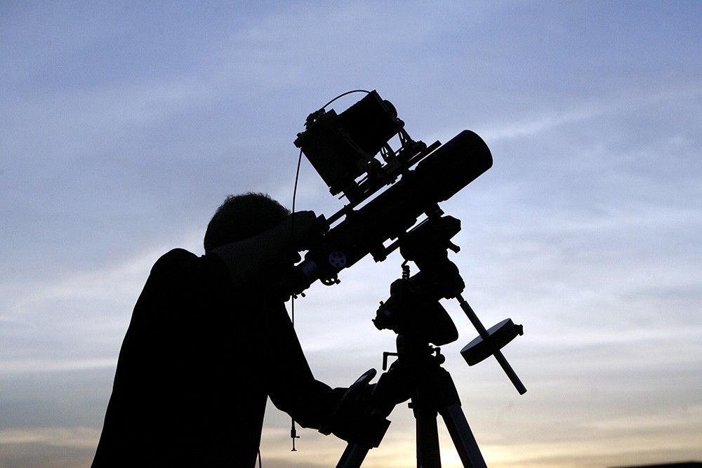 A man with a telescope is silhouetted against a blue sky