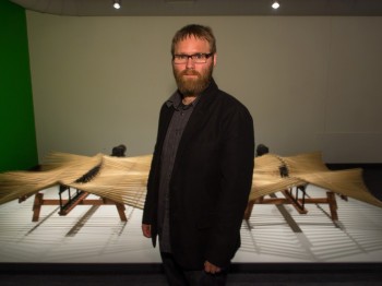 A man stands in front of a large wooden object with patterns of undulating wooden sticks