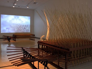 An art gallery with two large objects made of many wooden rods and spikes and a projection of clouds on the wall
