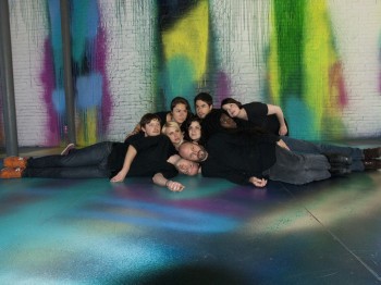 Nine people pose lying on top of each other in a room with colorful paint on the walls and floor.