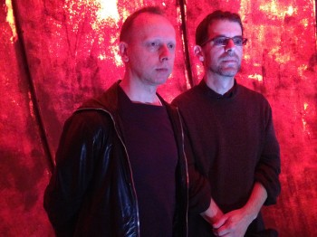 Two men stands against a non-uniform red background