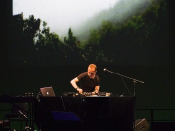 A man with a sound board, microphone, and laptop performs on a stage in front of a projected image of a forest