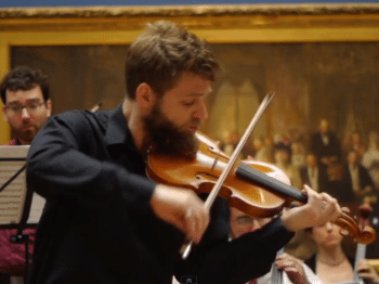 Two men perform violin in front of a large oil painting