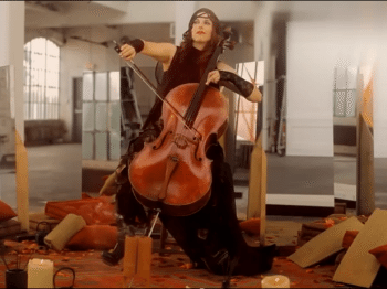 Video will of a woman playing cello surrounded by candles, pillows, and other objects
