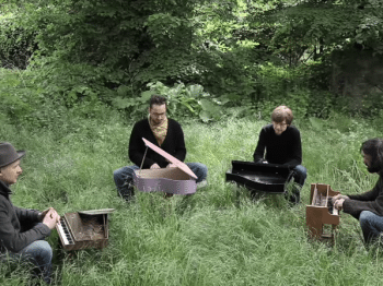 Four men play tiny pianos in a grassy field.