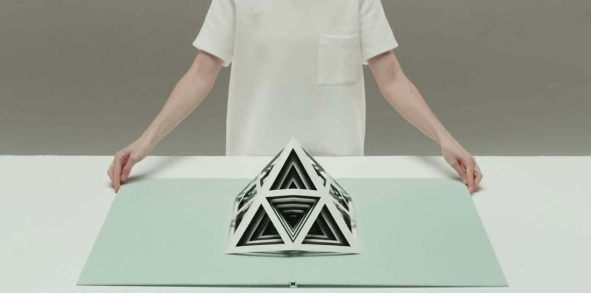 A person stands behind a pyramid-shaped sculpture made of triangles cut out from paper.