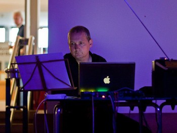 A man sits behind a laptop and a music stand.