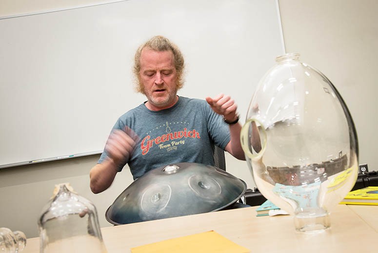 A man plays a metal drum next to a table with glass objects on it