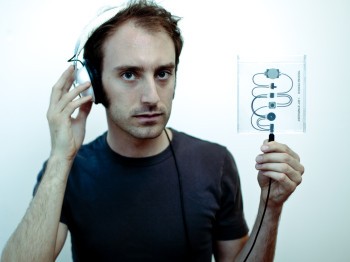 A man wearing headphones holds up a transparent plastic square with chips and wires running through it