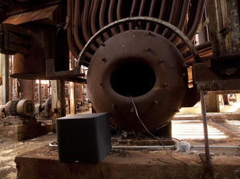 A modern sound speaker in front of large rusted machines