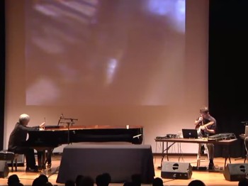 Two men on a stage, with a grand piano, electric guitar, multiple laptops, and a projection behind them.