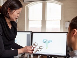 A professor points out an aspect of a flower-like object, a rendering of which is also shown on a computer screen.