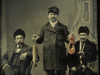 Historic photo of three men in a room. Two are sitting and one is standing, holding a fish and a lobster.