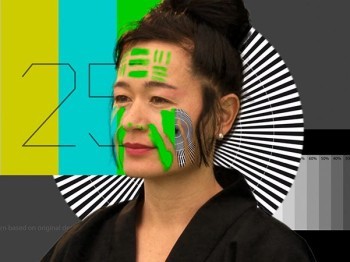 A woman with green, black, and white images on her face stands in front of graphics patterns in color and greyscale.