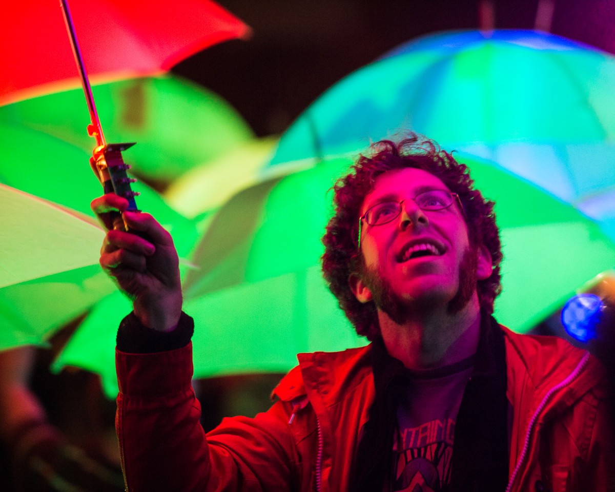 A participant in the Umbrella Project stands under an umbrella among a crowd of multicolored umbrellas lit from within by LEDs..