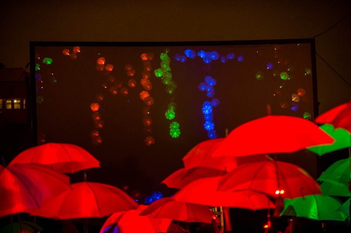 A crowd of multicolored umbrellas lit from within by LEDs are projected onto a screen in the background.