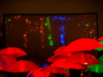 A crowd of multicolored umbrellas lit from within by LEDs are projected onto a screen in the background.